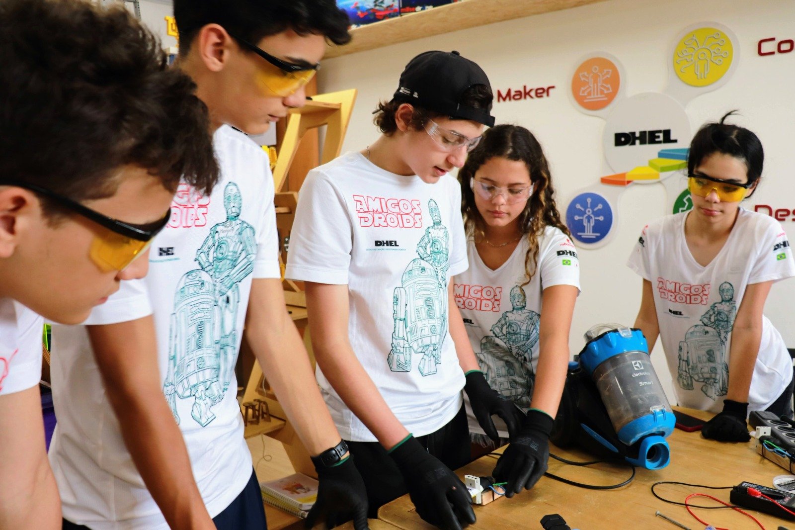 Estudantes da equipe Amigos Droids, da DHEL, com óculos de proteção, trabalhando em eletrônica na bancada maker